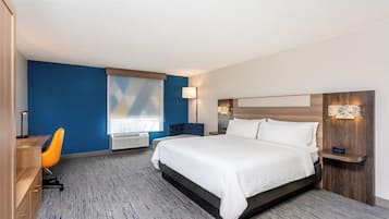 A hotel room with a bed, a desk, a chair, a window with blinds, and a wall-mounted lamp.