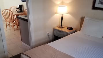 Una habitación de hotel con una cama, una mesita de noche con lámpara, una pequeña mesa con cafetera y una silla.