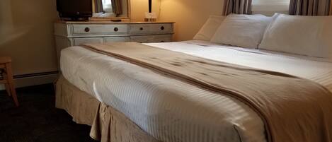 A neatly made bed with a beige comforter, a bedside lamp, a television on a dresser, and a window with curtains.