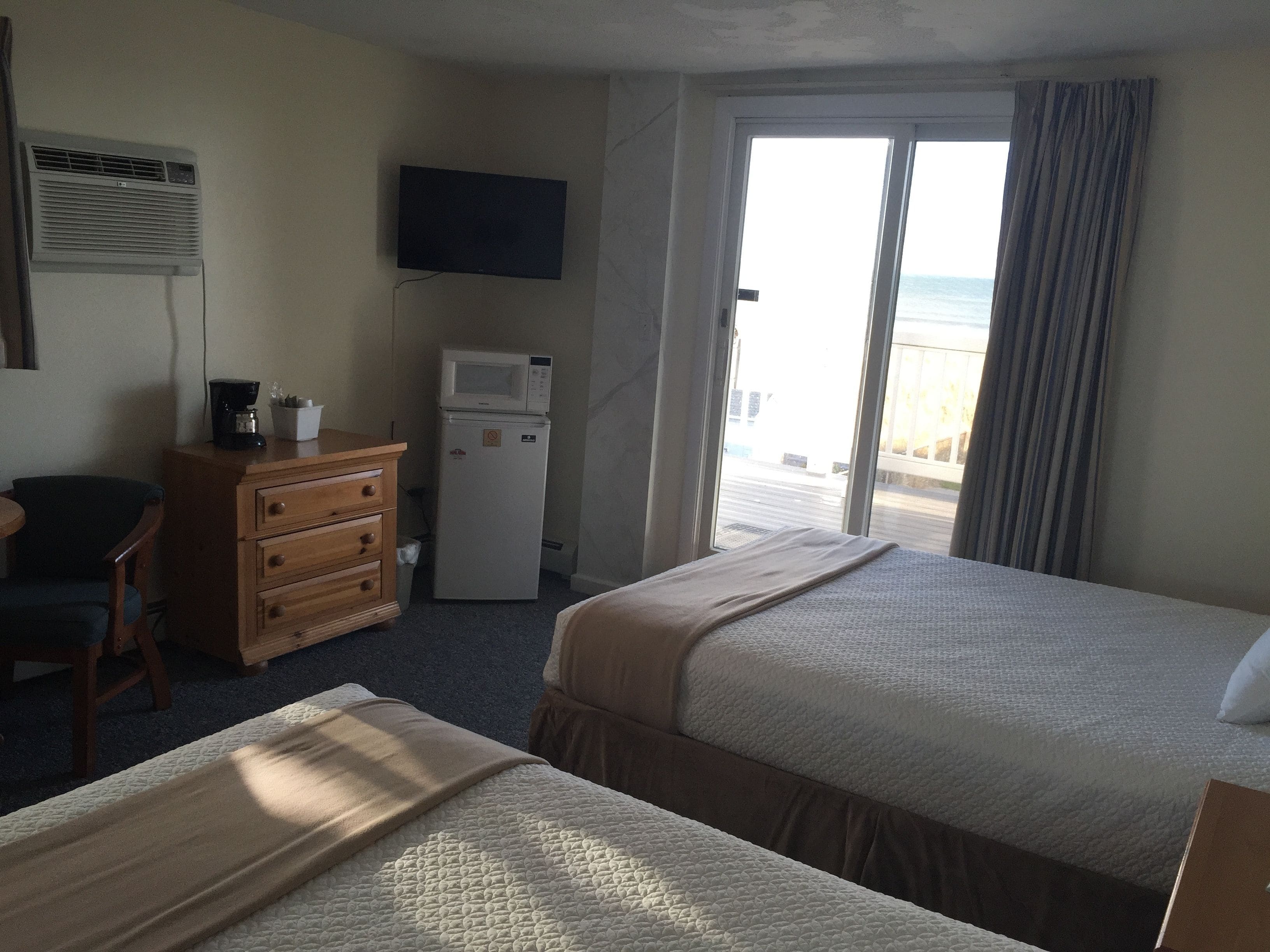 A hotel room with two beds, a TV, a microwave, a coffee maker, a dresser, and a view of the beach.