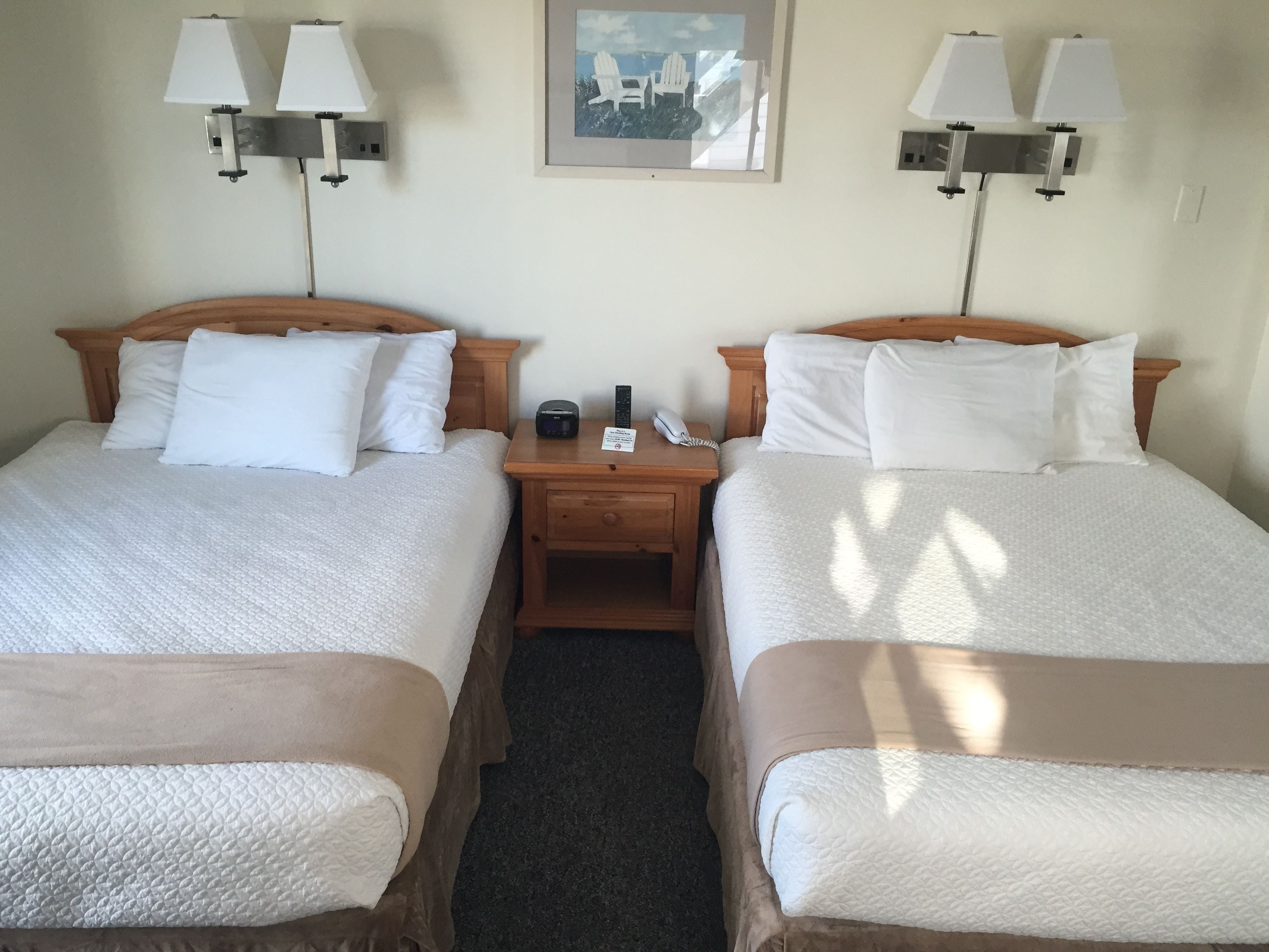 A hotel room with two beds, each with white bedding and a wooden headboard. There are two bedside tables, one on each bed, with lamps and a phone on them. A framed picture hangs on the wall above the beds.
