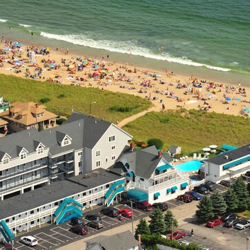 A coastal hotel with multiple floors, a parking lot, and a beach in the background.