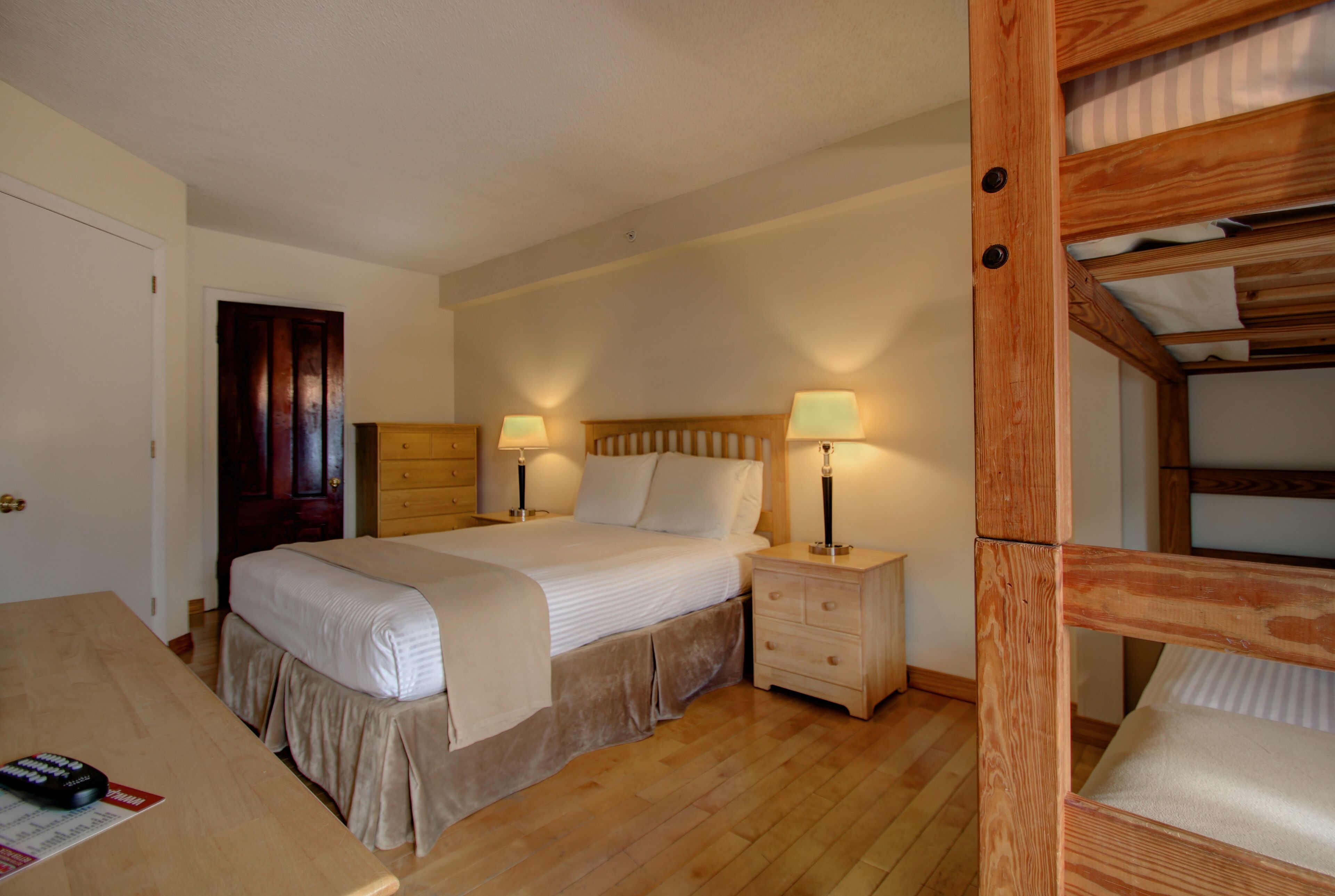 A bedroom with a wooden bunk bed, a single bed, a bedside table with lamps, a wooden dresser, and a wooden desk with a remote control.