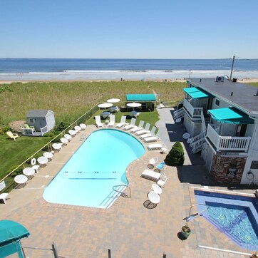 A swimming pool with lounge chairs and umbrellas, a separate hot tub, and a view of the beach.
