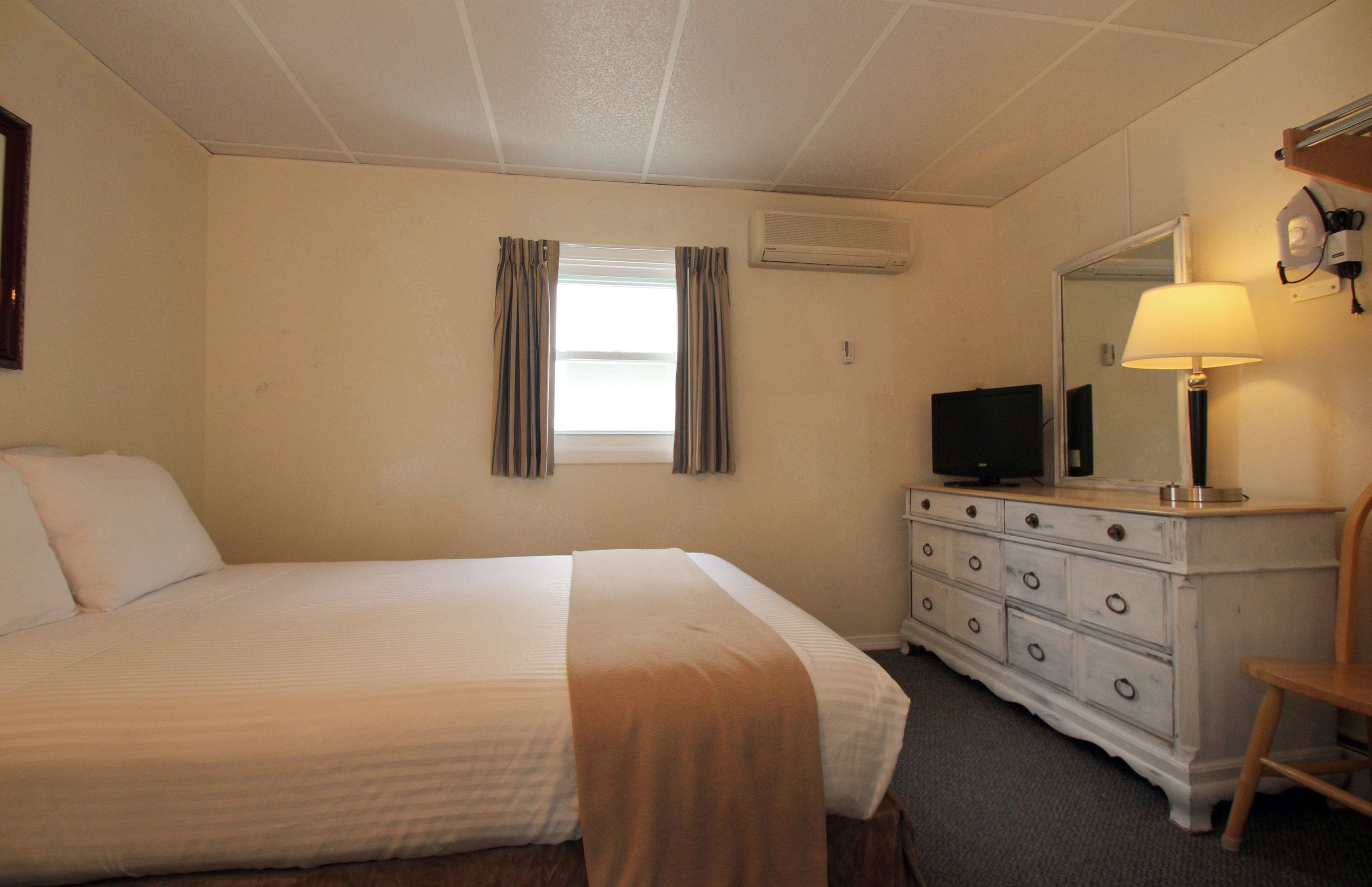 A hotel room with a bed, a dresser, a television, a lamp, and a window with curtains.