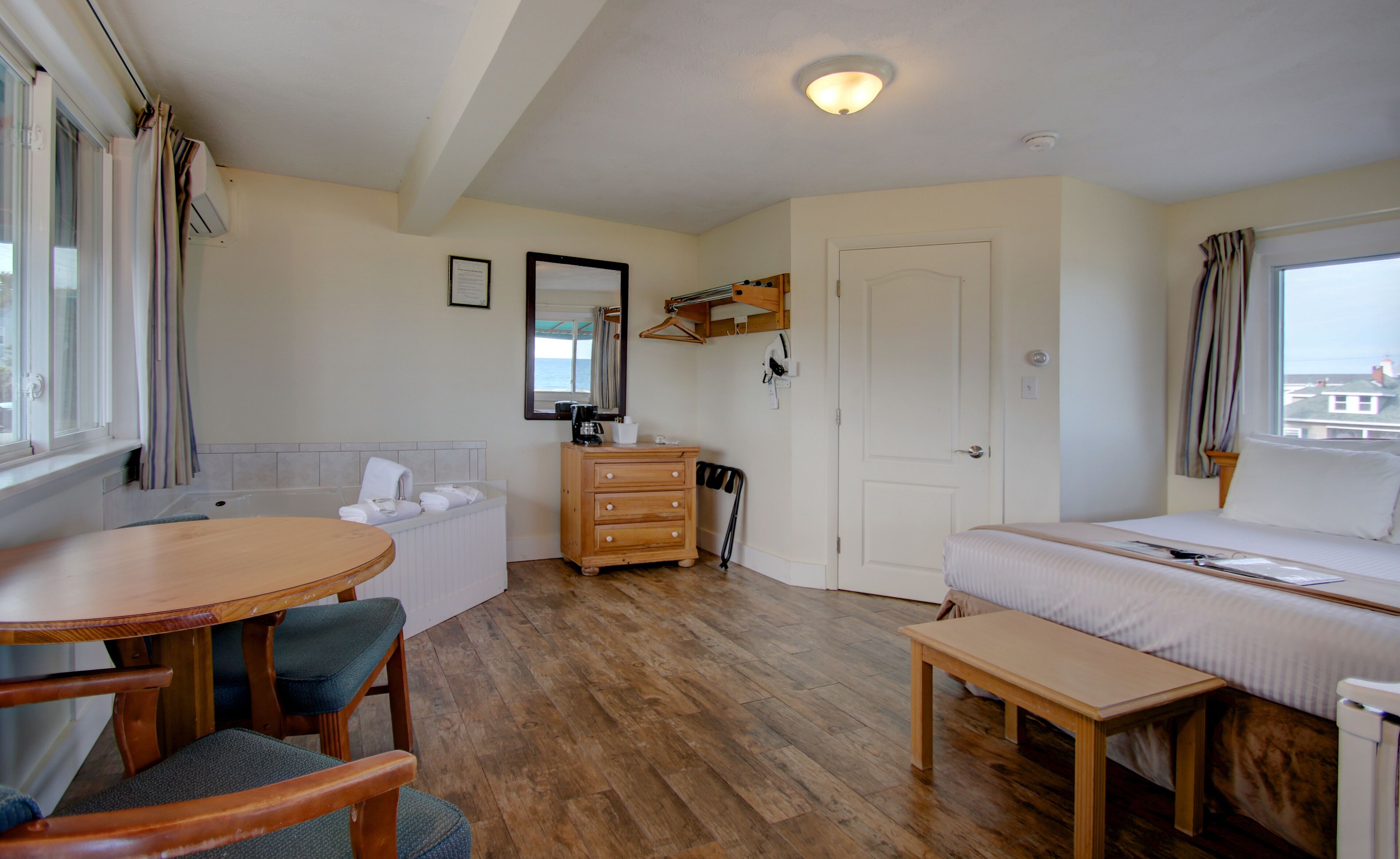 Habitación de hotel con suelo de madera, una cama, un escritorio con una silla, una mesita pequeña, un baño con bañera y una ventana con cortinas.