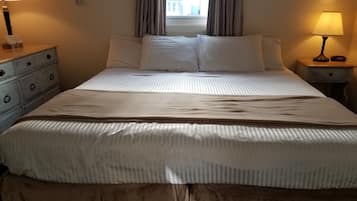 Una cama bien hecha con sábanas blancas, una mesita de noche de madera con una lámpara y una ventana con cortinas.