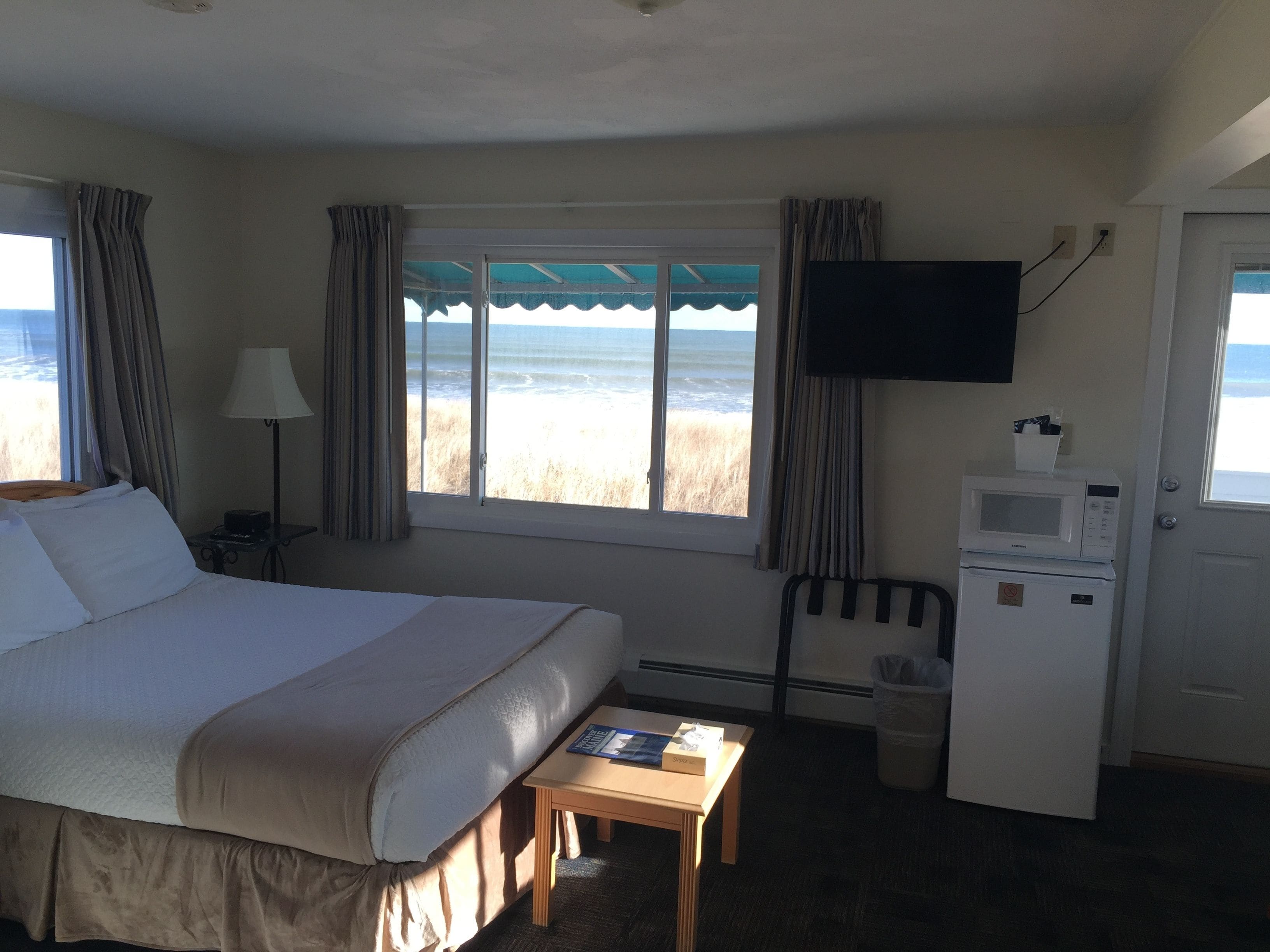 A hotel room with a bed, a desk, a television, a microwave, and a view of the beach.