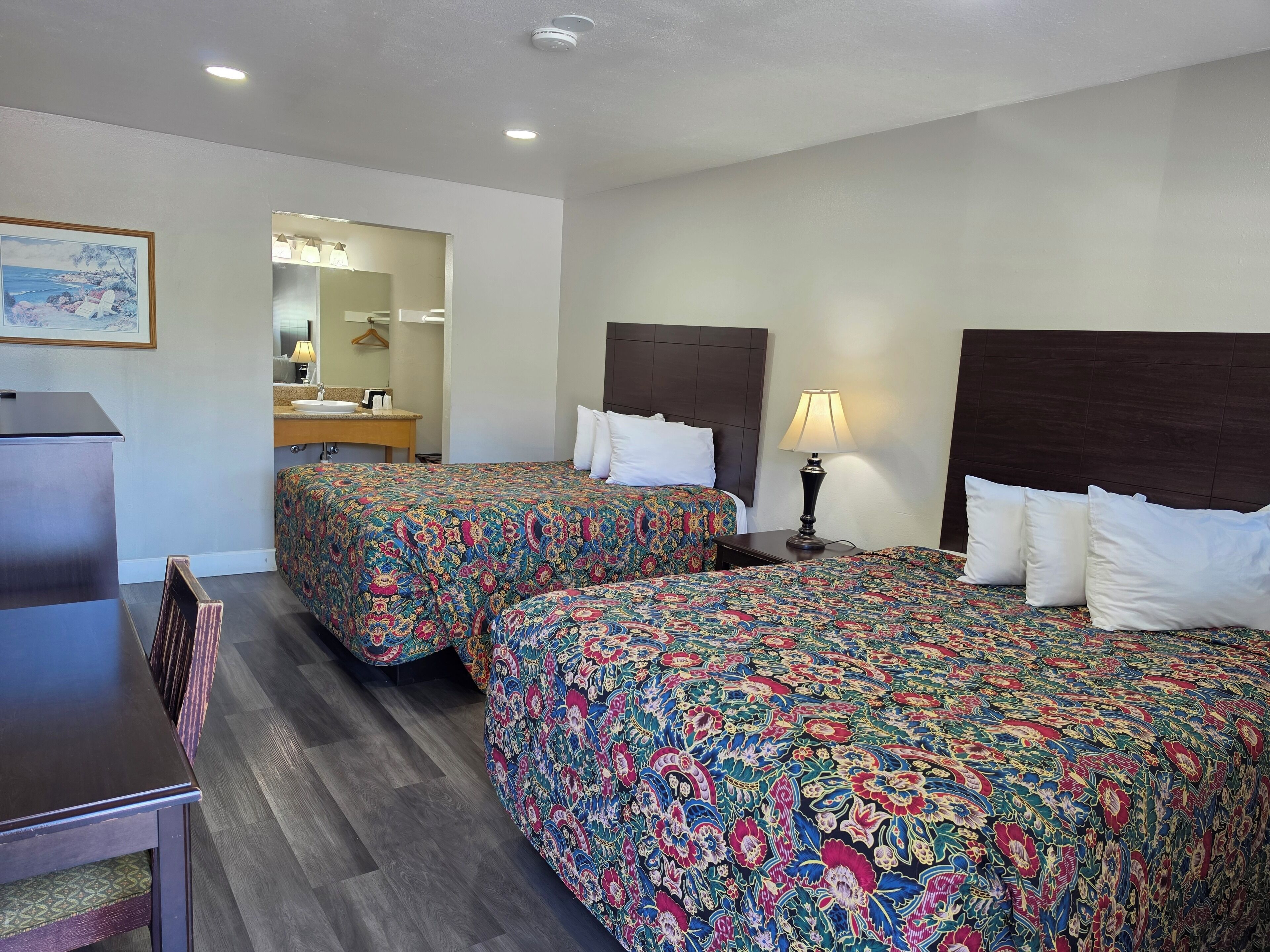 A hotel room with two beds, a desk, a chair, a mirror, and a lamp.