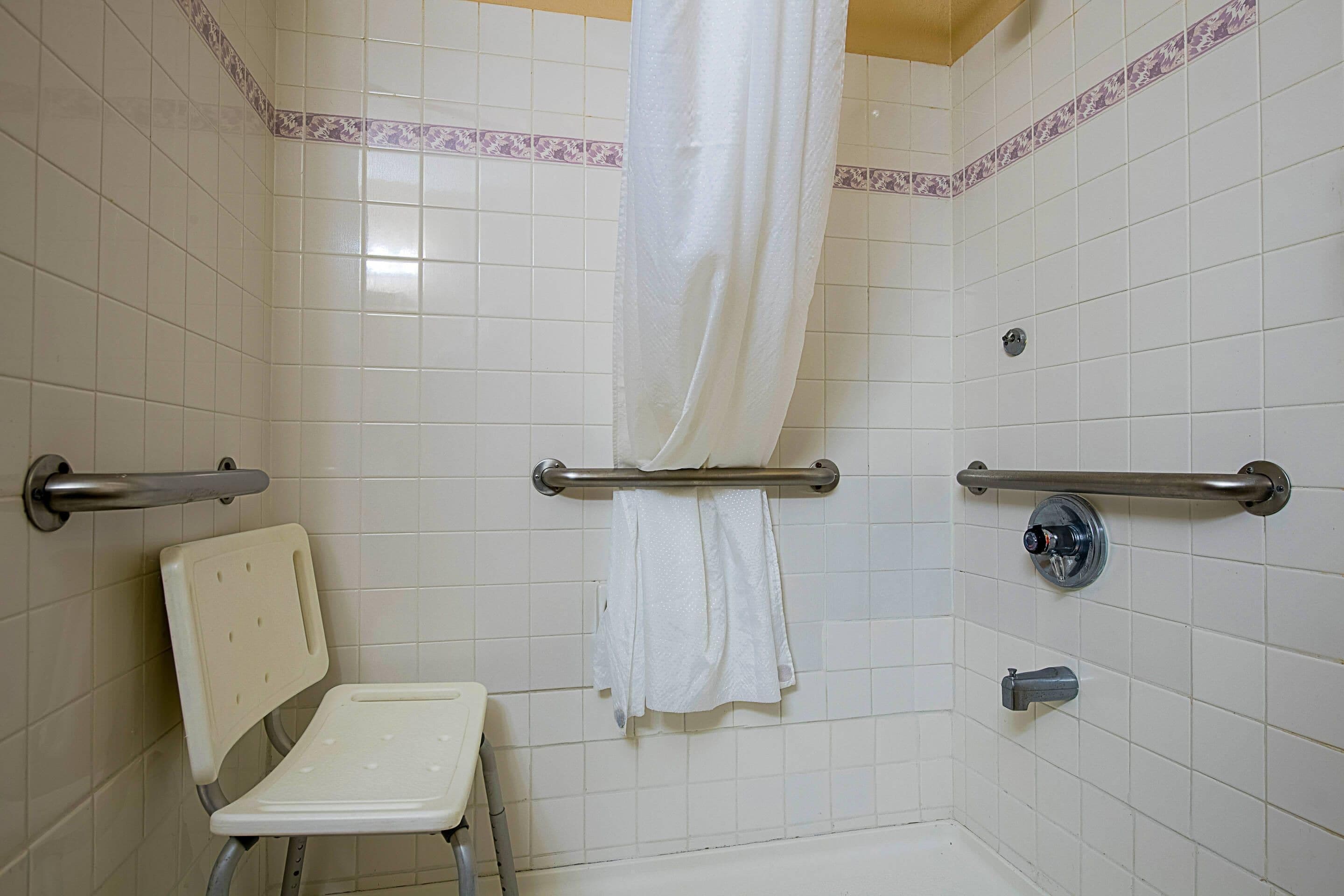 A bathroom with white tiles, a shower with a curtain, a grab bar, a white chair, and a towel rack.