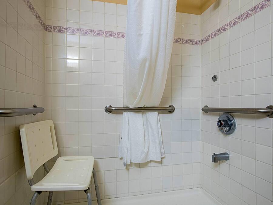 Un baño con azulejos blancos, una ducha con cortina, un asidero, una silla blanca y un perchero para toallas.