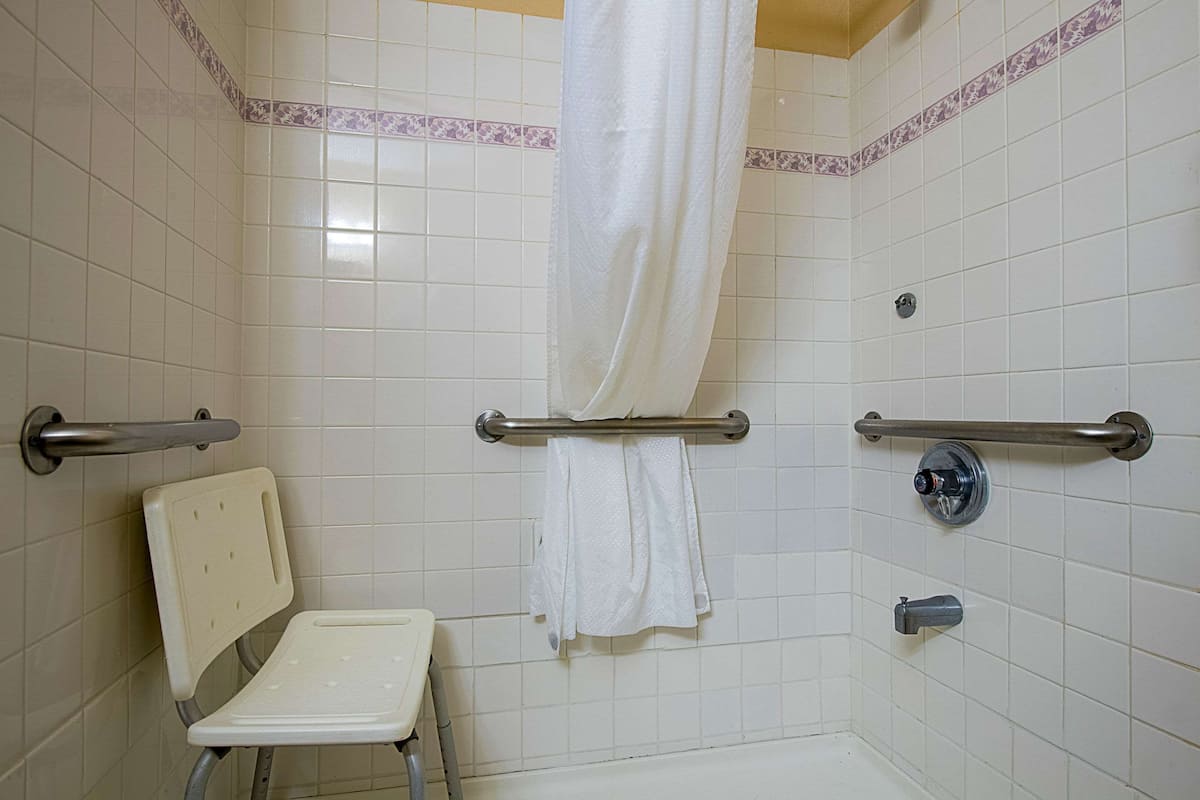 Une salle de bain avec des murs carrelés de blanc, une douche avec une porte en verre, une barre d’appui, une chaise blanche et un porte-serviettes.