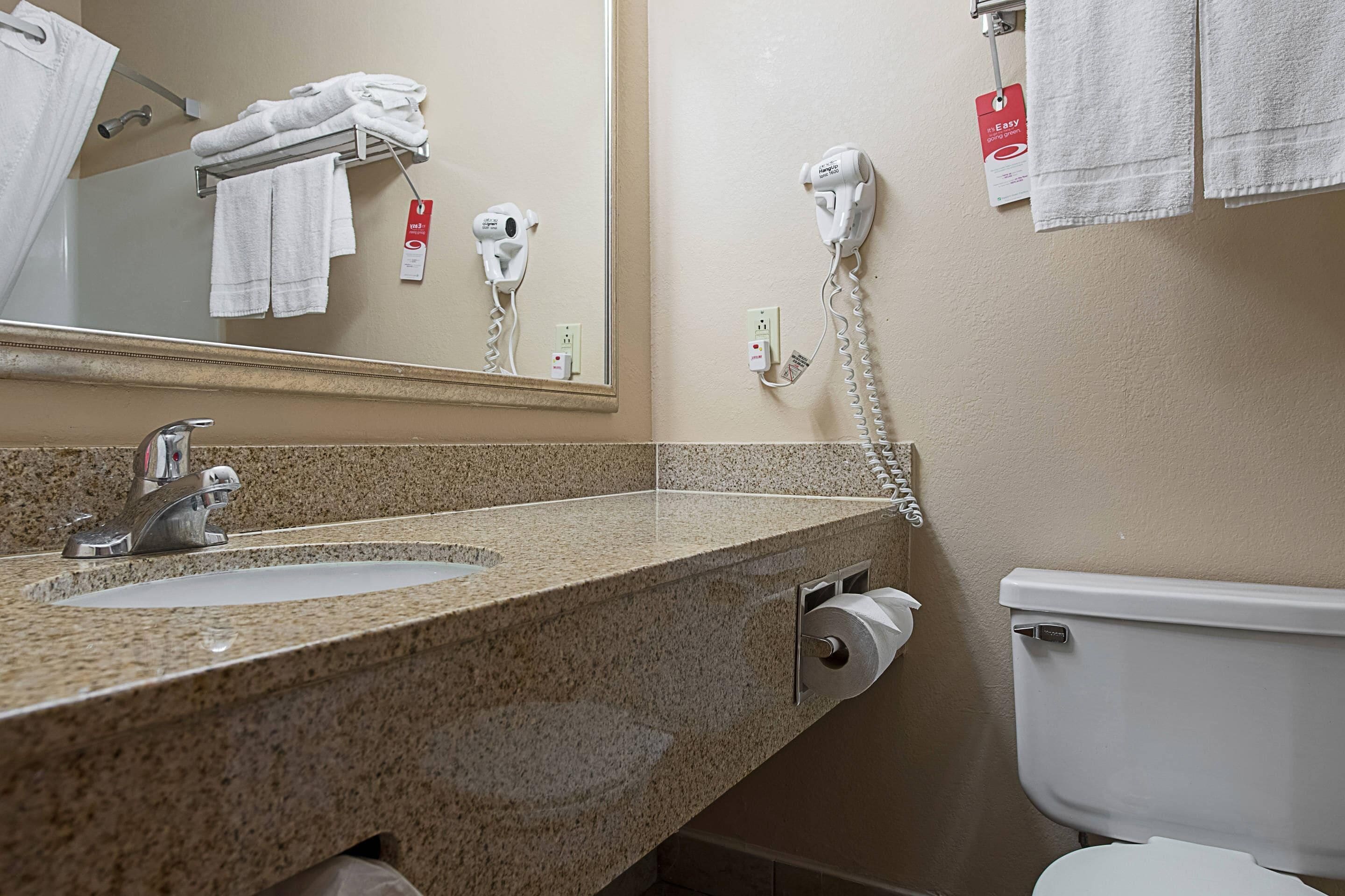 Un baño con encimera de granito, un lavabo, un espejo, un inodoro y un secador de pelo en un soporte de pared.