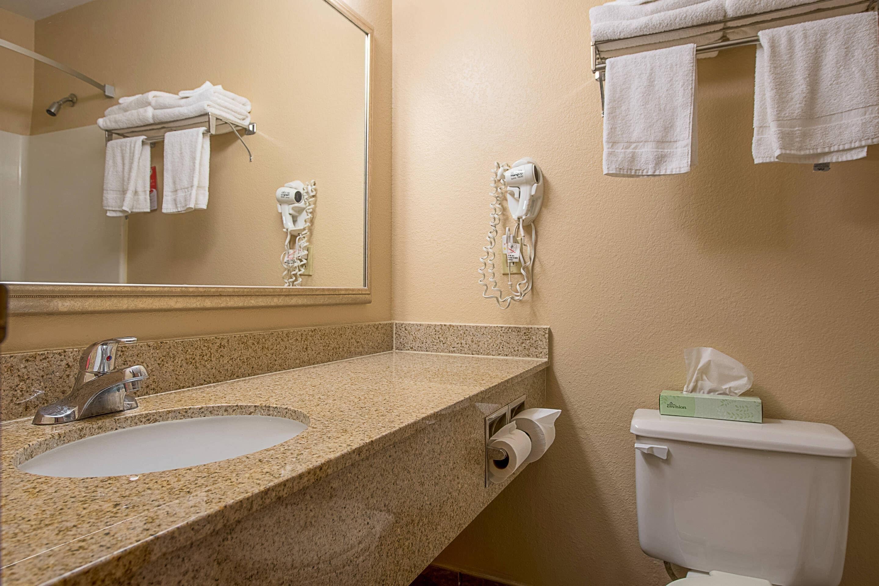 A bathroom with a granite countertop sink, a mirror, a towel rack with folded towels, a hairdryer, and a toilet with a tissue box.
