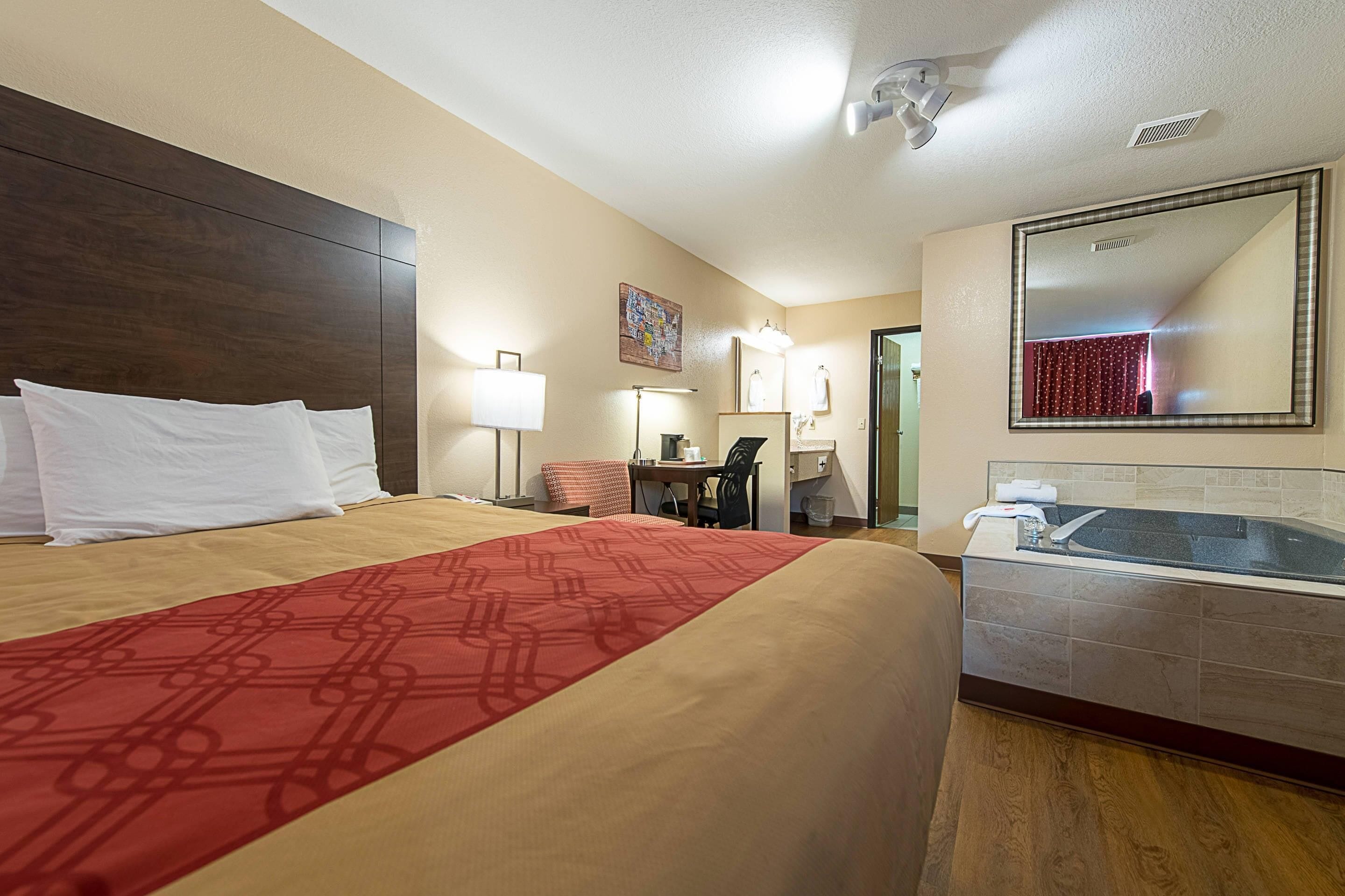 Habitación de hotel con una cama grande, un escritorio con silla, una lámpara de escritorio, un espejo montado en la pared y una bañera.