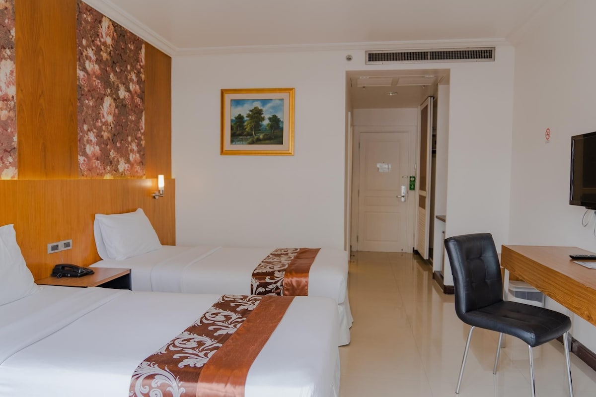 Quarto de hotel com duas camas, uma mesa, uma cadeira, televisão, pintura e porta que dá para outro cômodo.