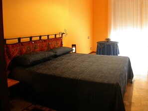 A bedroom with a bed, a nightstand, a small table, and a window with blinds.