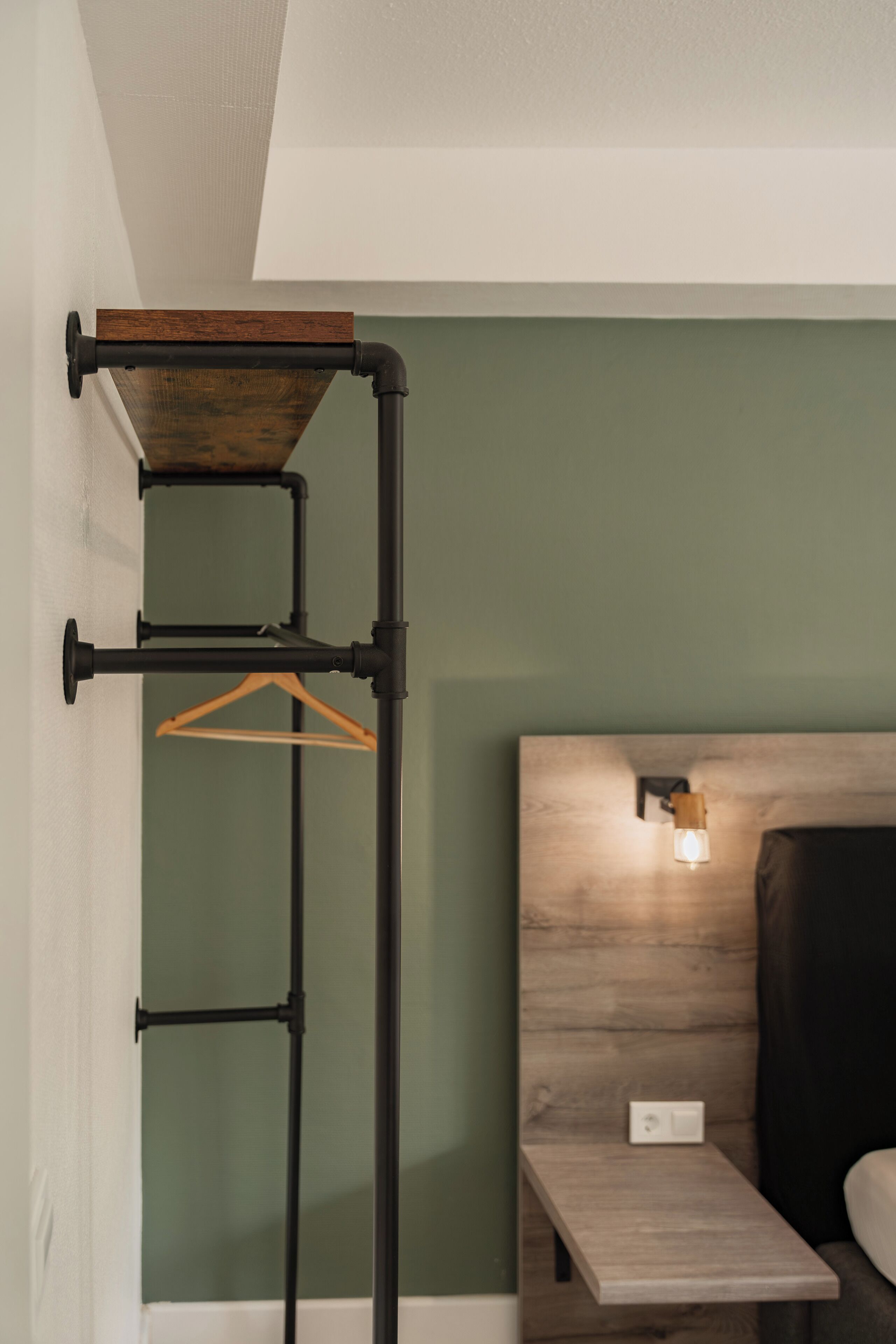 Una habitación con cama, cabecero de madera, mesita de noche y estante fijado a la pared con un perchero de madera.