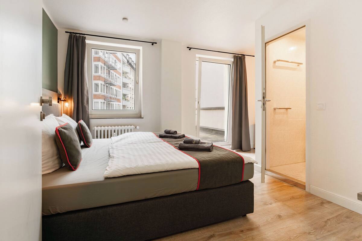 A modern bedroom with a large bed, a window with curtains, a view of buildings, and a door leading to a balcony.