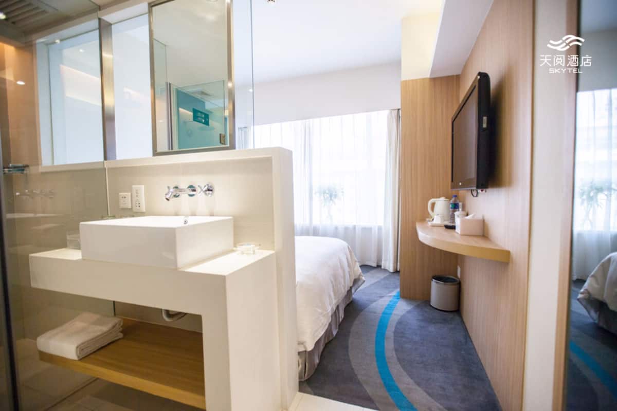 A modern hotel room with a large white sink, a mirror, a flat-screen TV mounted on the wall, a bed with white bedding, and a small shelf with toiletries.