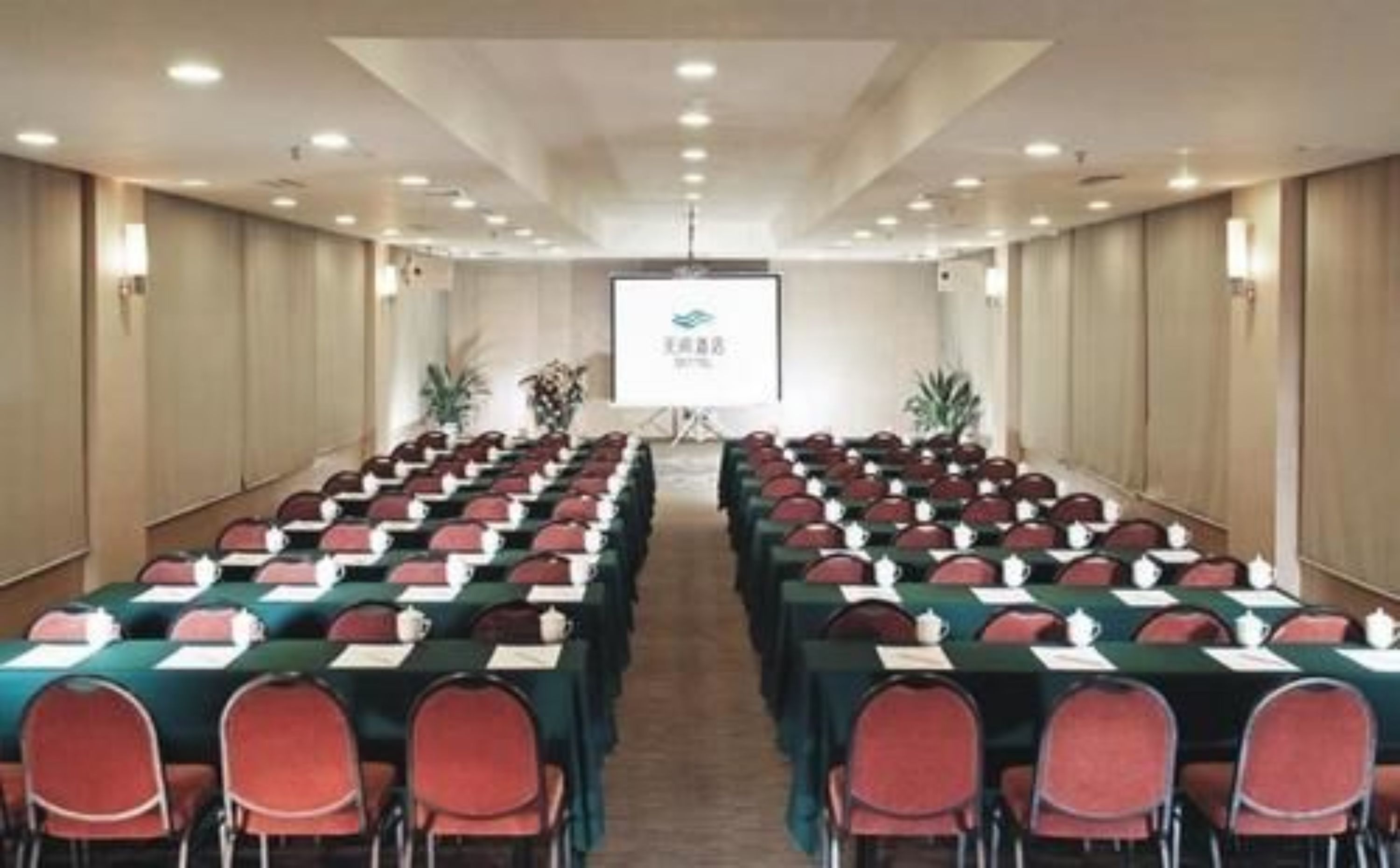 A conference room with rows of tables and chairs, a projector screen at the front, and a logo on the screen.