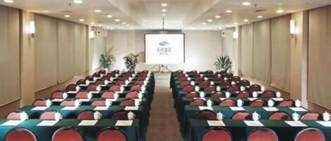 A conference room with rows of tables and chairs, a projector screen at the front, and a logo on the screen.