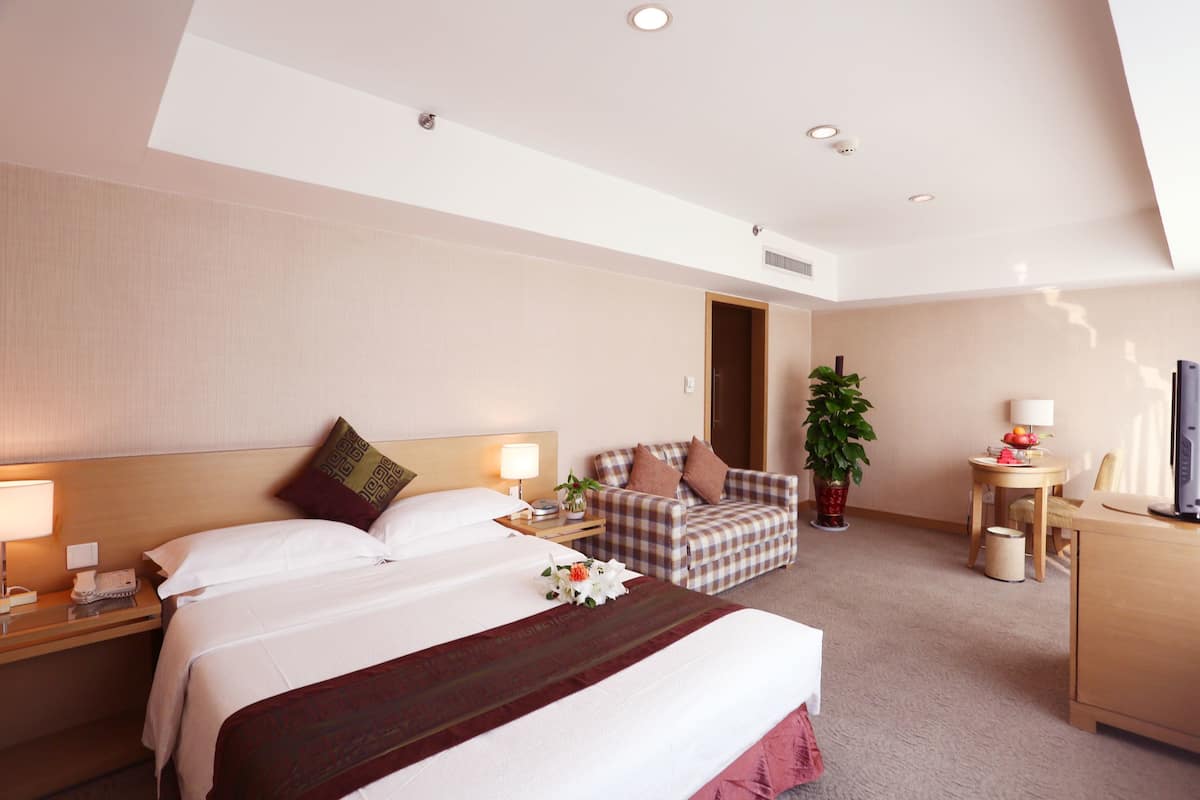 A hotel room with a large bed, a desk, a chair, a small table with a lamp, and a potted plant.