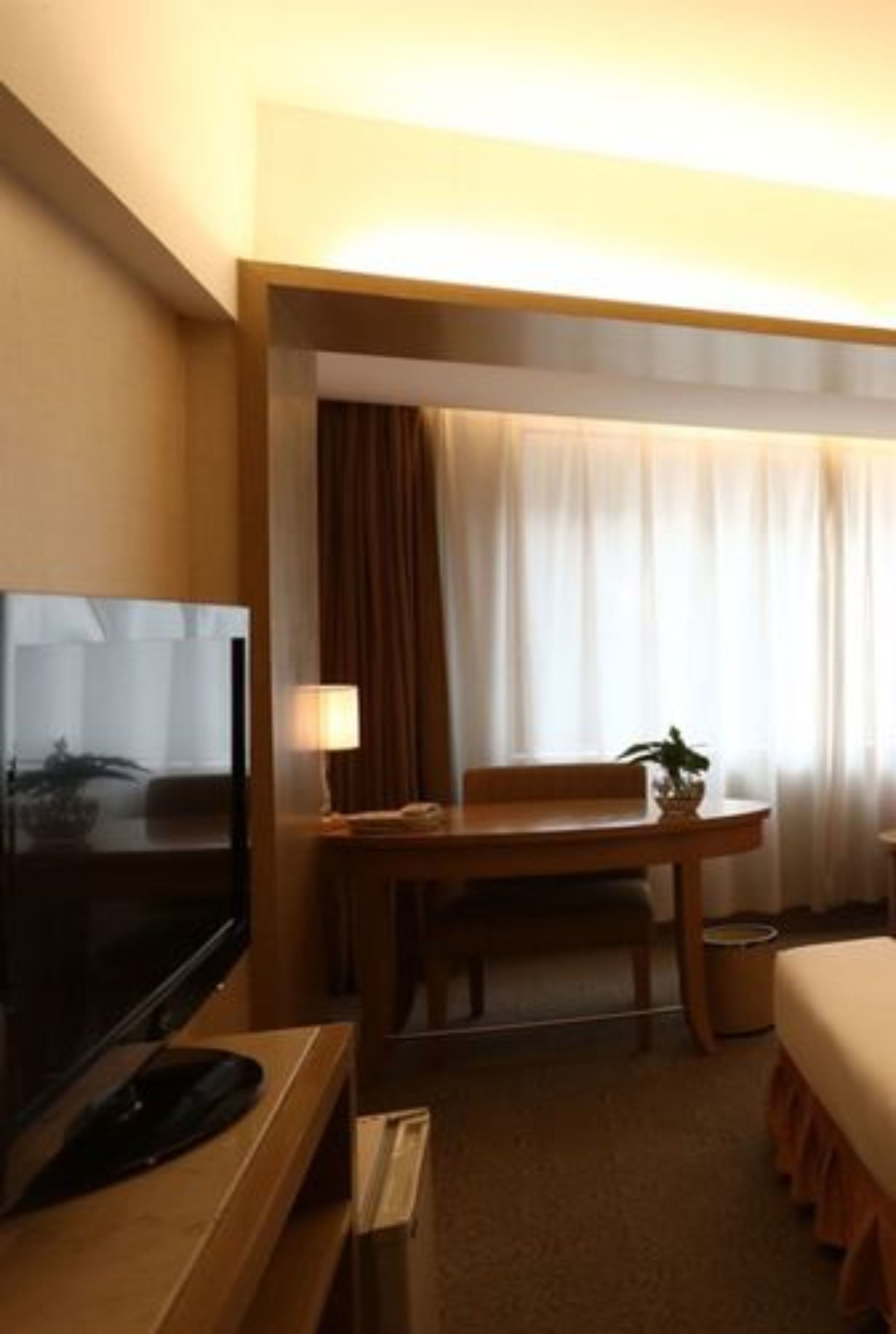 Ein Hotelzimmer mit einem Bett, einem Schreibtisch mit Computer, einem Fernseher, einer Lampe und einem Fenster mit Vorhängen.
