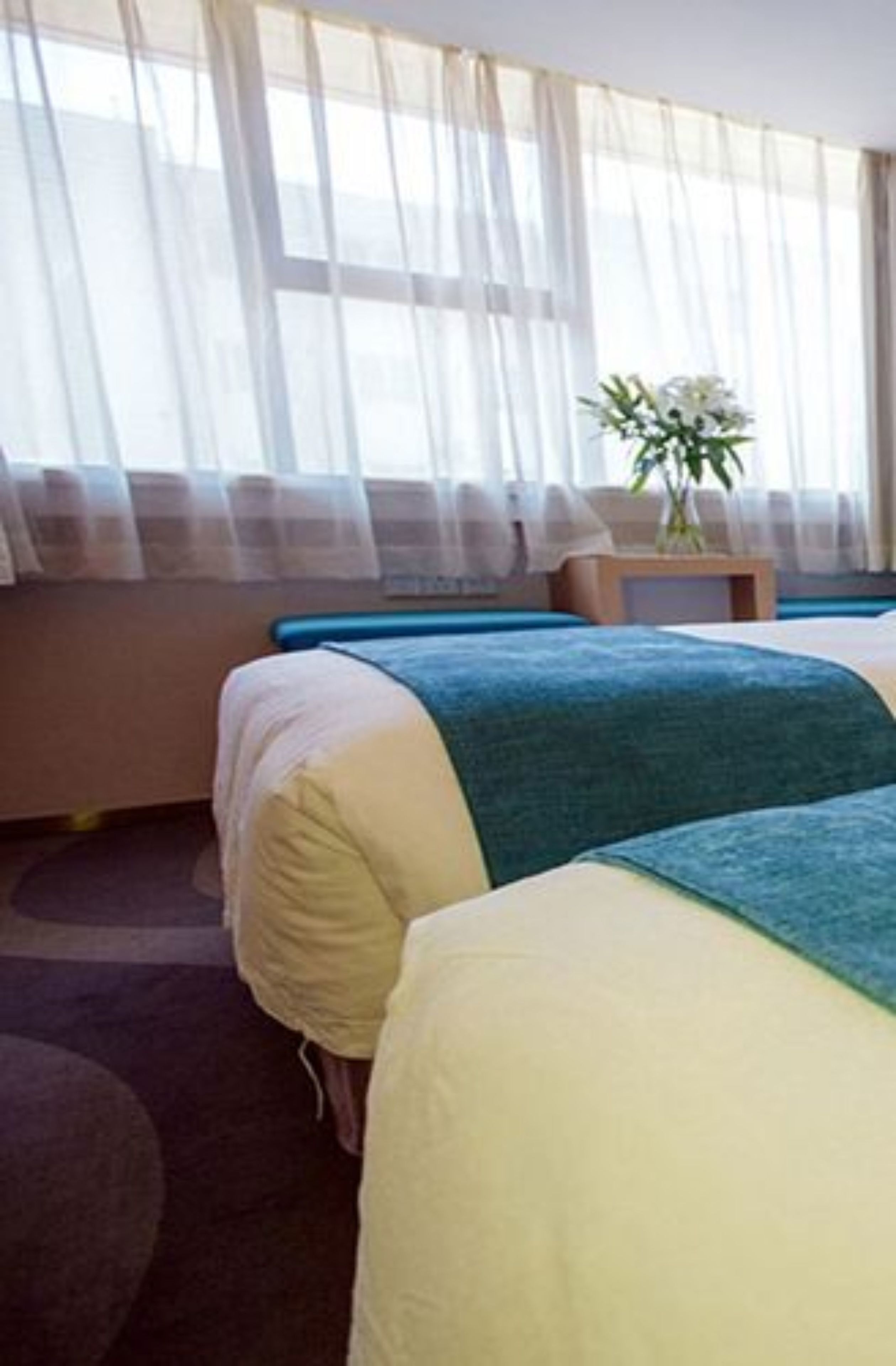 Una habitación de hotel con dos camas, una mesita con un florero y cortinas de encaje.