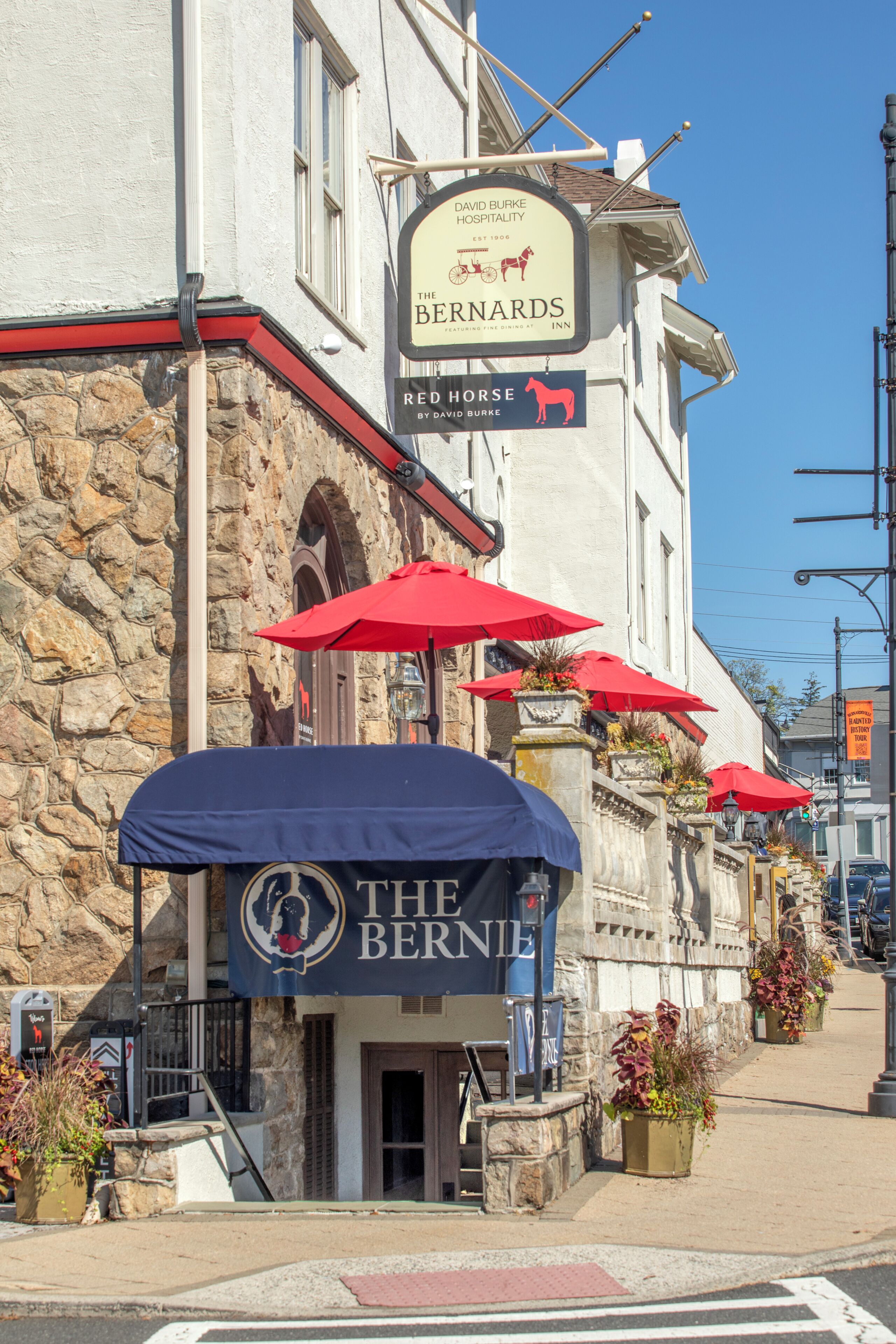 A building with a sign for 'The Bernards' featuring a horse logo, a stone facade, and red umbrellas.