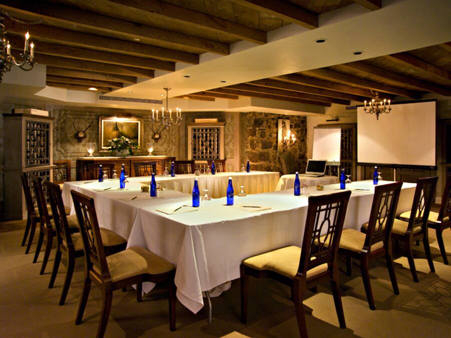 A long dining table with chairs, a chandelier, and a whiteboard in a room with stone walls and wooden beams.