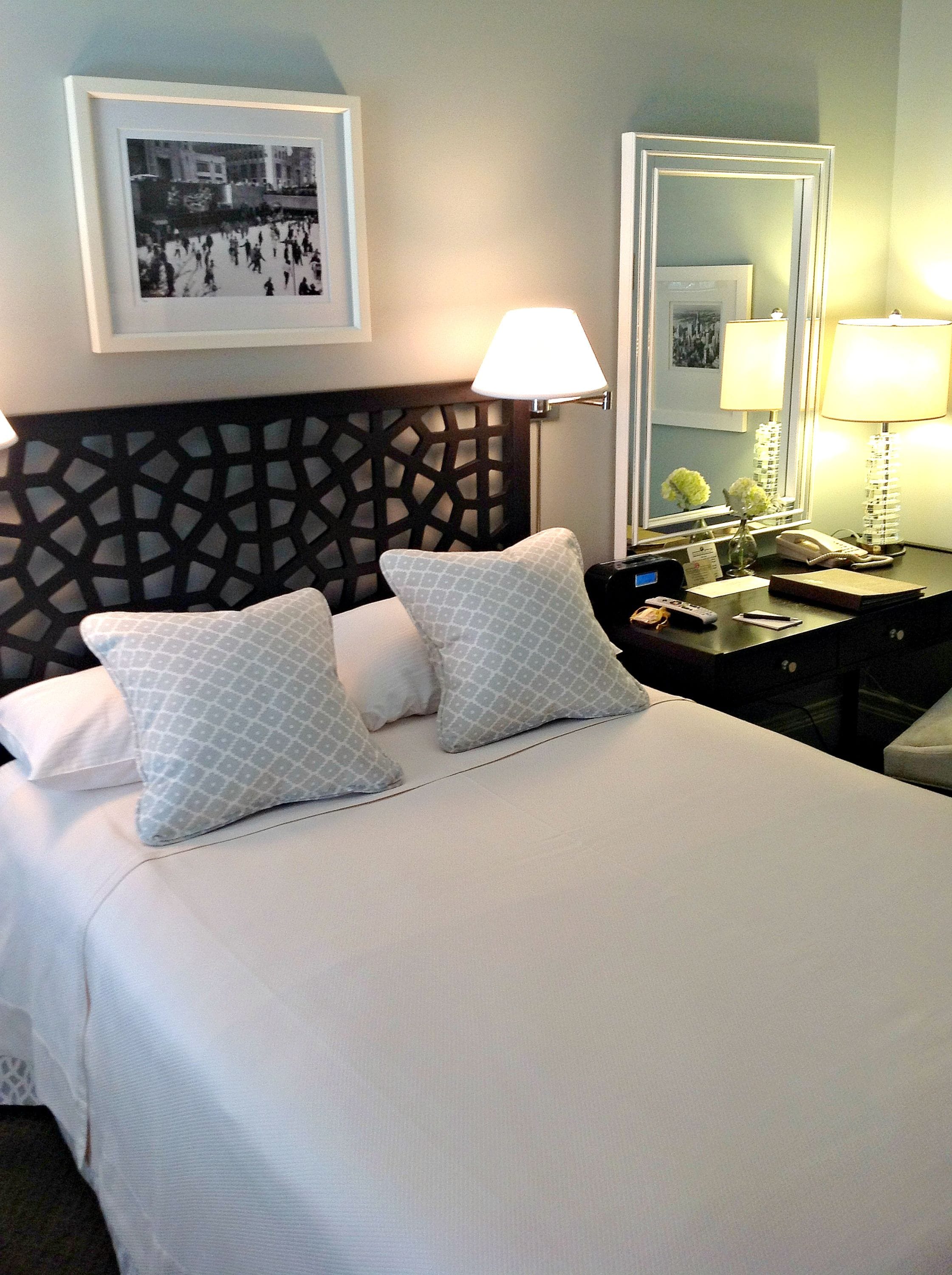 A hotel room with a neatly made bed, two pillows, a headboard with a geometric pattern, a bedside table with a lamp, a mirror, and a framed picture on the wall.