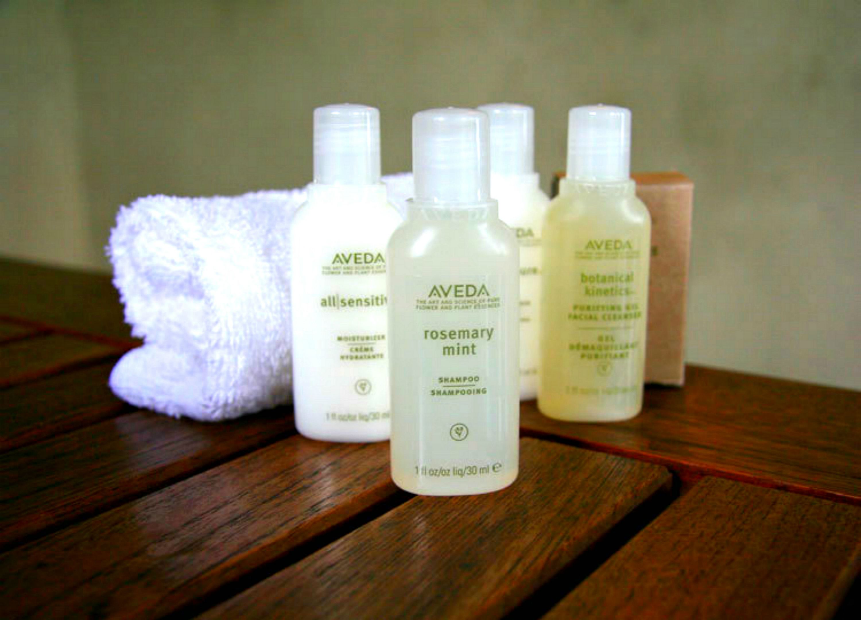 Superficie de madera con varios productos de cuidado de la piel Aveda, incluyendo una botella de champú romero y menta, junto con otros artículos.