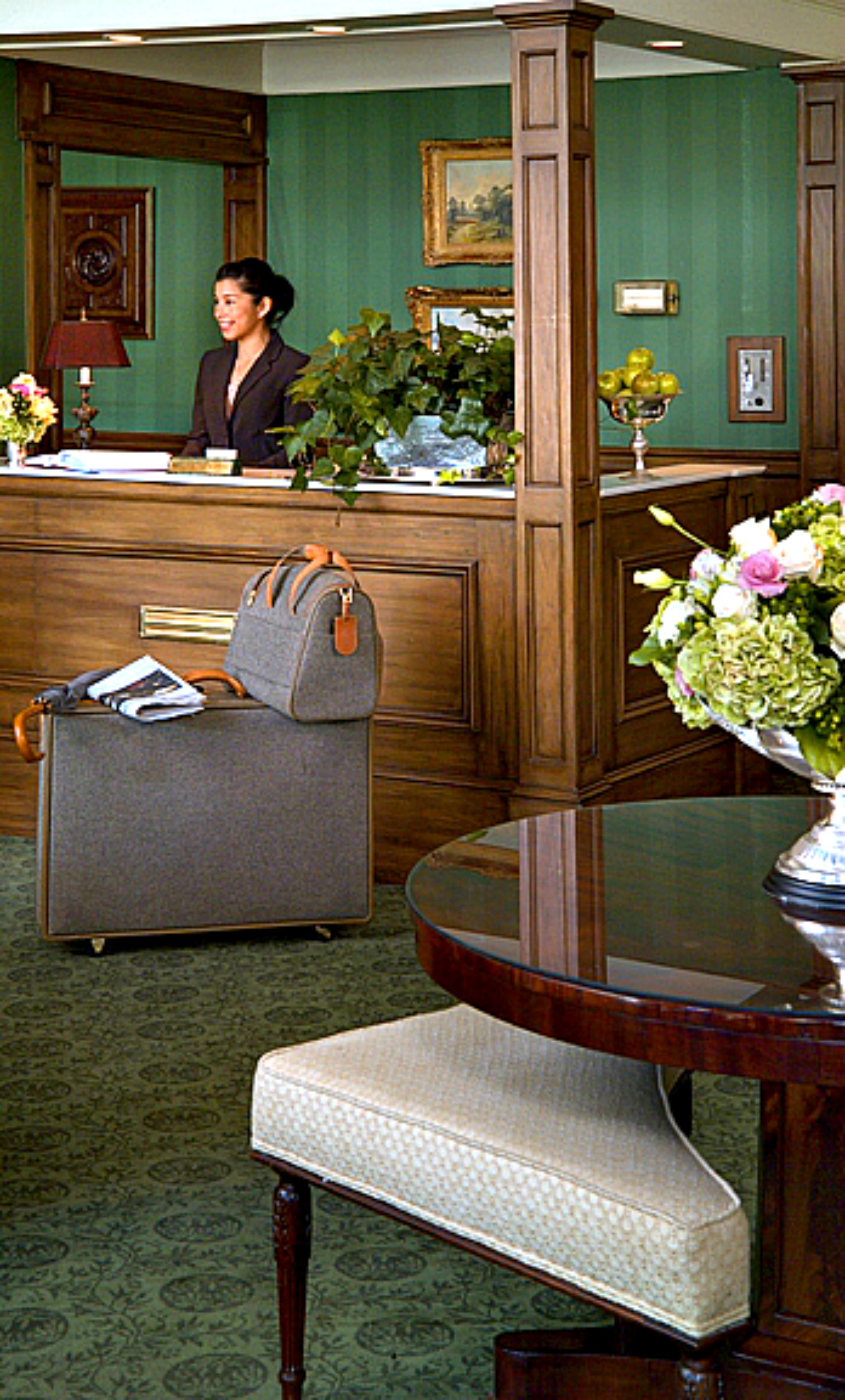 A reception area with a receptionist, a desk, a grey armchair, a round table with a vase of flowers, and a patterned carpet.