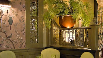 A dining area with a table set for two, a potted plant, and a decorative wall with a floral pattern.