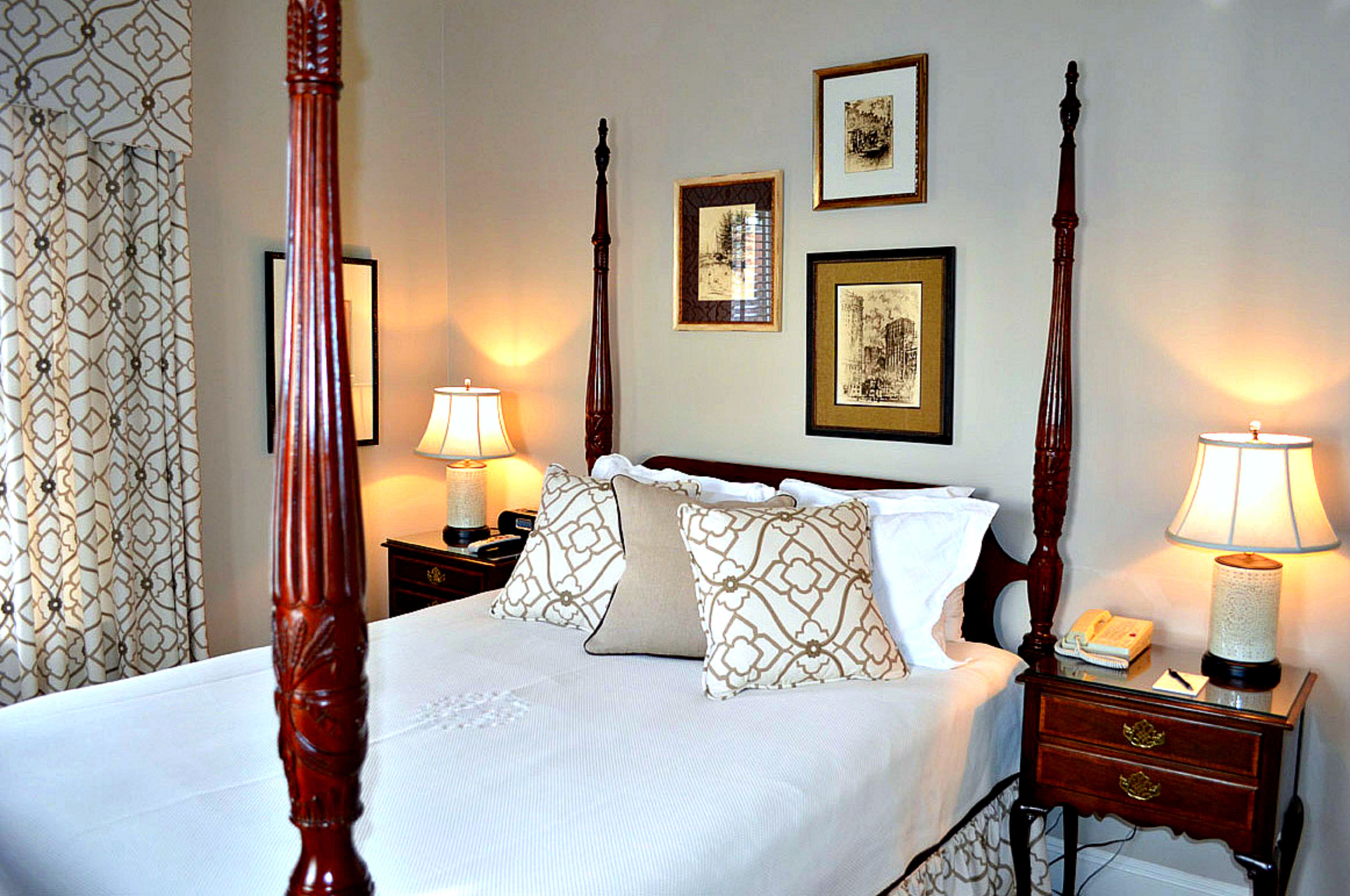 A four-poster bed with a white bedspread and patterned pillows, flanked by two bedside tables with lamps, a nightstand with a telephone, and framed pictures on the wall.