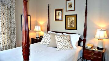 A four-poster bed with a white bedspread and patterned pillows, flanked by two bedside tables with lamps, a nightstand with a telephone, and framed pictures on the wall.