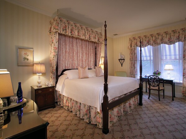 A four-poster bed with floral curtains, a bedside table with a lamp, and a chair by the window.