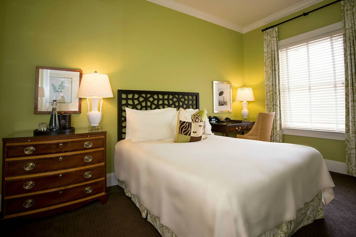 A neatly made bed with a headboard, a dresser with drawers, two bedside lamps, and a window with curtains.