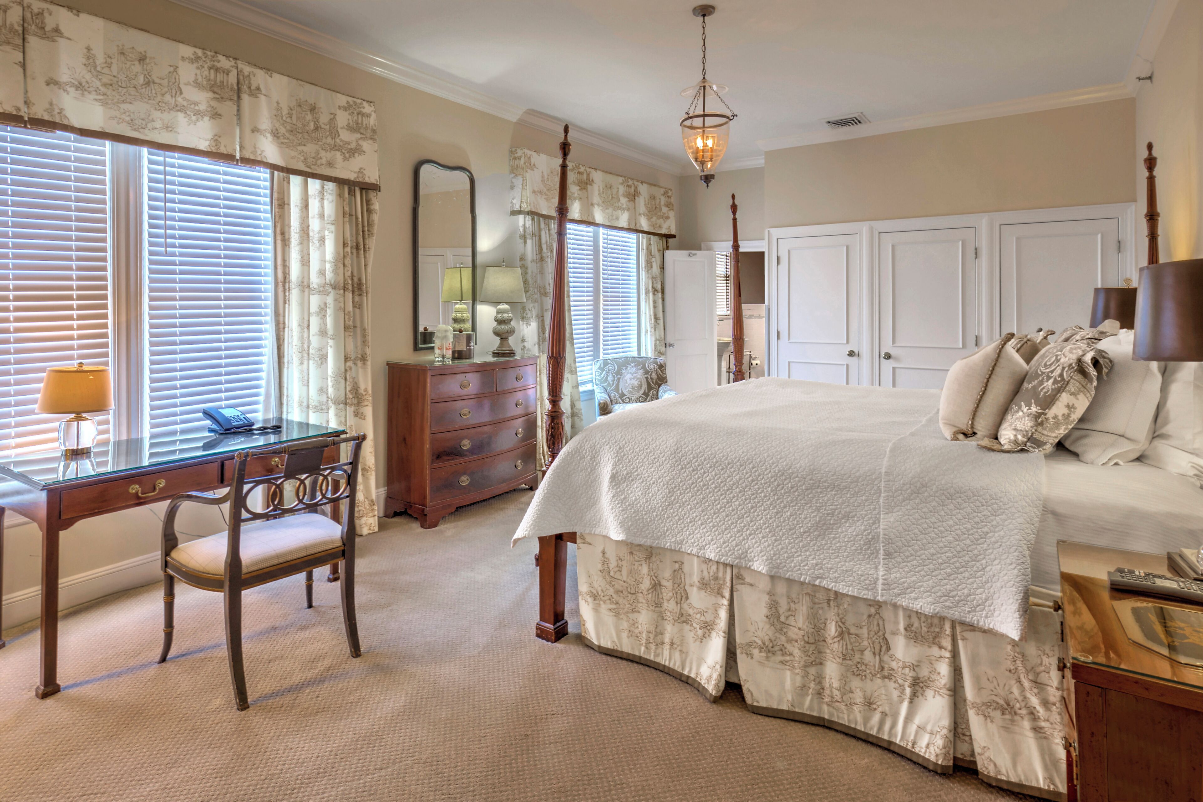 A bedroom with a four-poster bed, a desk with a lamp, a dresser, and a large mirror.