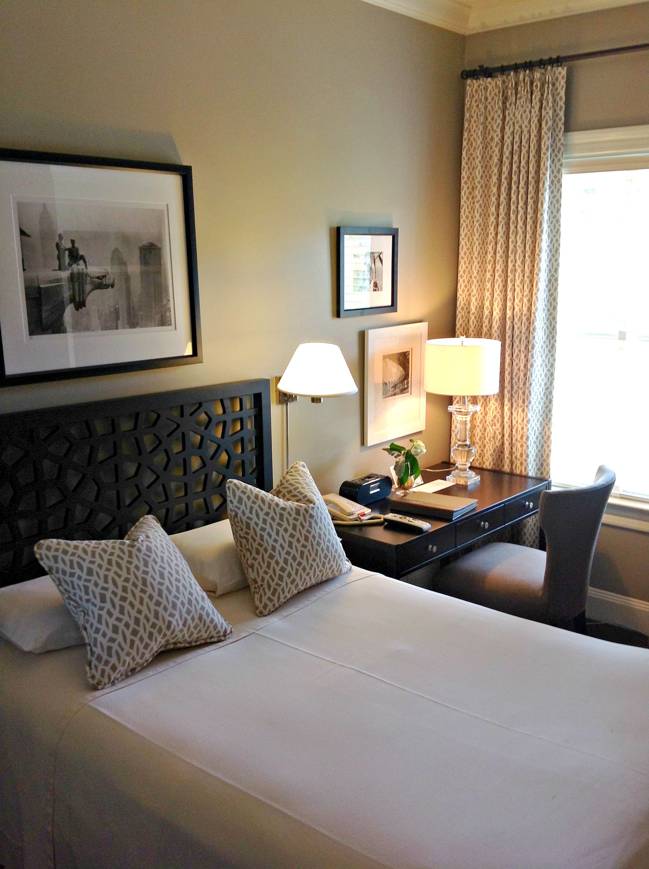 A hotel room with a bed, two pillows, a desk with a lamp, a telephone, a remote control, and framed pictures on the wall.