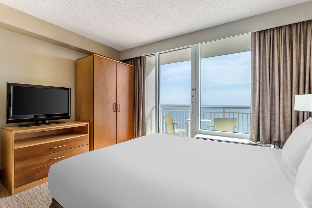 A hotel room with a large bed, a TV on a wooden cabinet, and a balcony with a view of the sea.