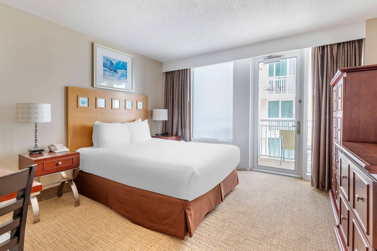 A hotel room with a large bed, a desk with a lamp, a chair, a wardrobe, and a balcony with curtains.
