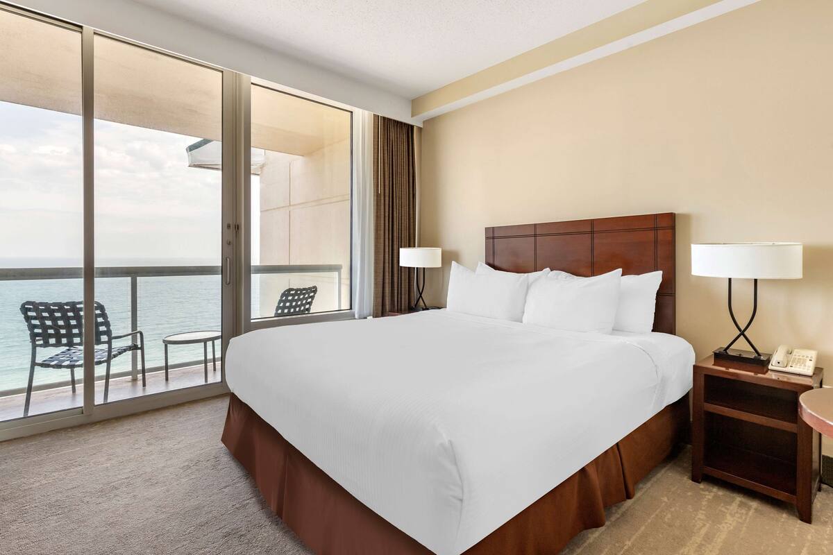 A hotel room with a large bed, a nightstand with a lamp, a chair by the window, and a view of the ocean.