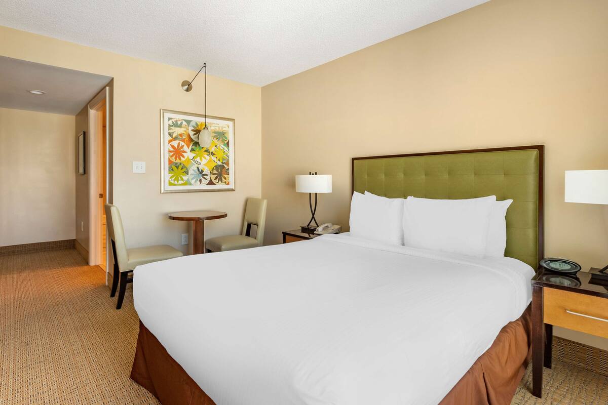 A hotel room with a large bed, a desk with a lamp, a chair, and a small table.