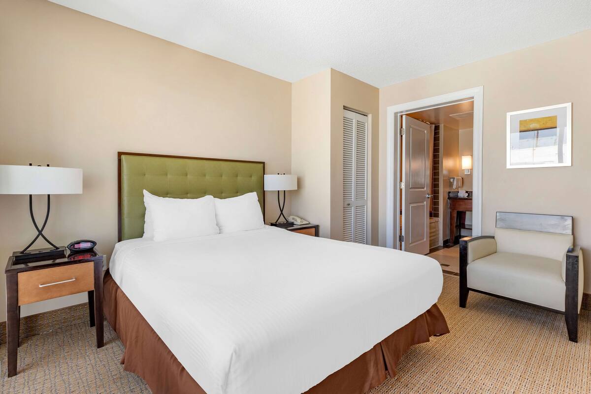 A hotel room with a large bed, bedside tables with lamps, a chair, a wardrobe, and a door leading to a bathroom.
