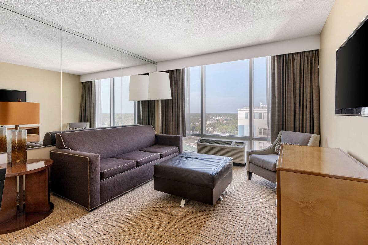 A modern hotel room with a large window, a sofa, a small table, and a desk.