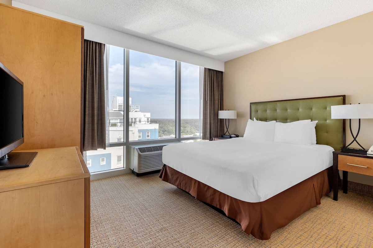A hotel room with a large bed, a television, a desk with a lamp, and a view of the city through large windows.