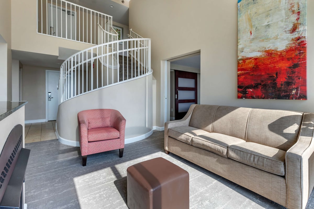 A modern living room with a beige sofa, a red armchair, a brown ottoman, a white spiral staircase, and a large abstract painting.