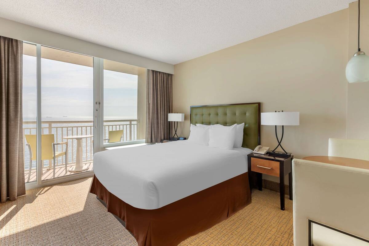 A hotel room with a large bed, a desk with a lamp, a chair, and a view of the ocean through a sliding door.